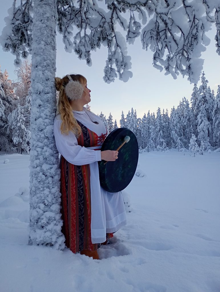 A Finnish singer in traditional dress holding a drum, looking towards a snowy forest — a meaningful experience in Finland through music and storytelling.